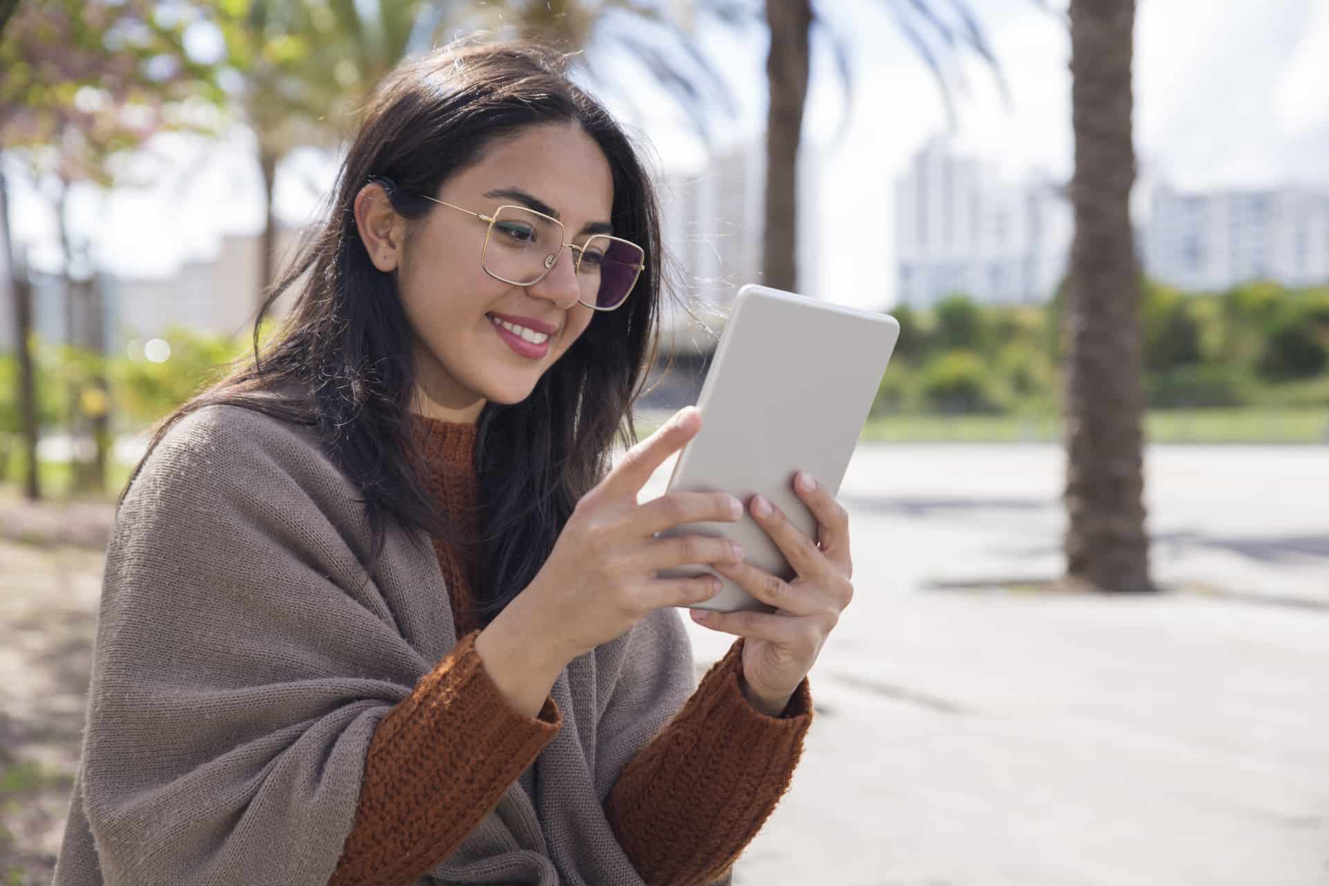 Smiling pretty woman using tablet computer outdoors. Young woman holding gadget with blurred trees and walkway in background. Technology concept.