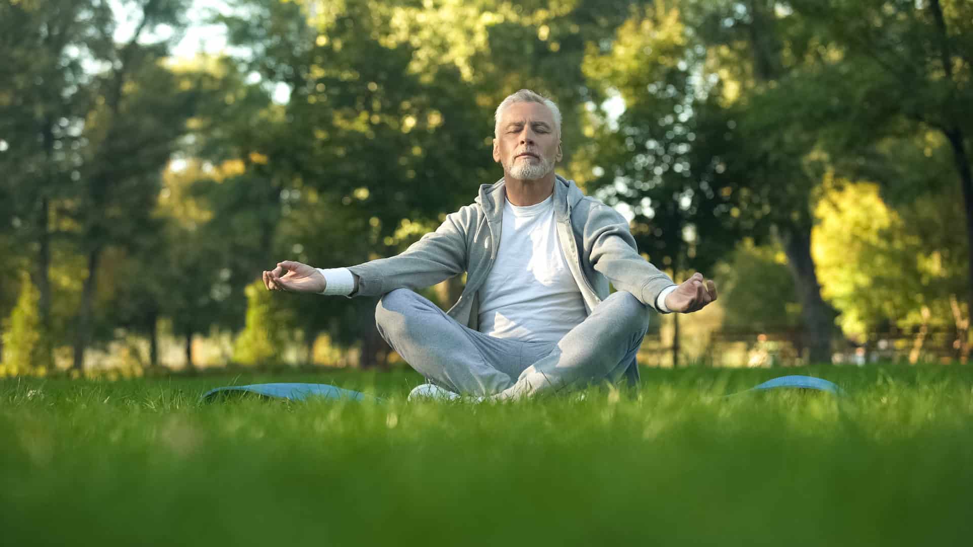 Healthy senior man meditating in park, sitting lotus pose on yoga mat, zen