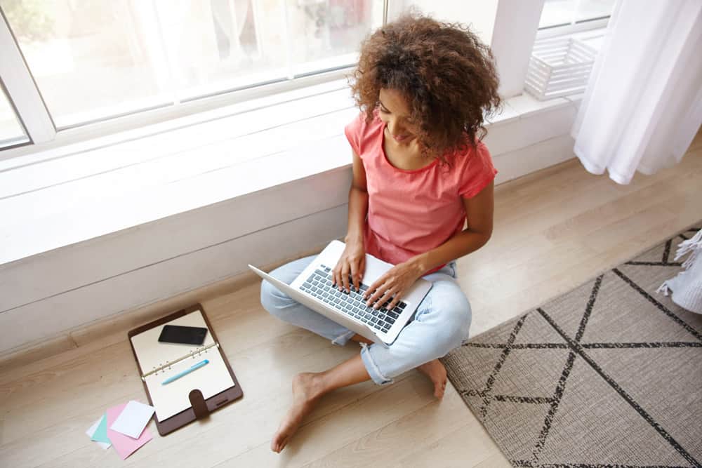 indoor-shot-young-dark-skinned-female-with-brown-curly-hair-sitting-floor-with-crossed-legs-writing-e-mail-with-her-modern-laptop