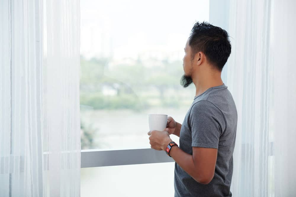 asian-man-standing-front-tall-window-with-mug-looking-out