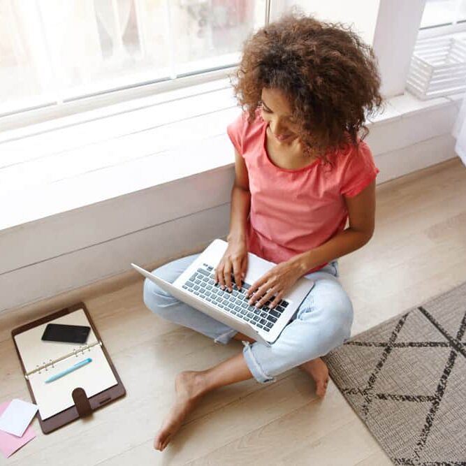 indoor-shot-young-dark-skinned-female-with-brown-curly-hair-sitting-floor-with-crossed-legs-writing-e-mail-with-her-modern-laptop